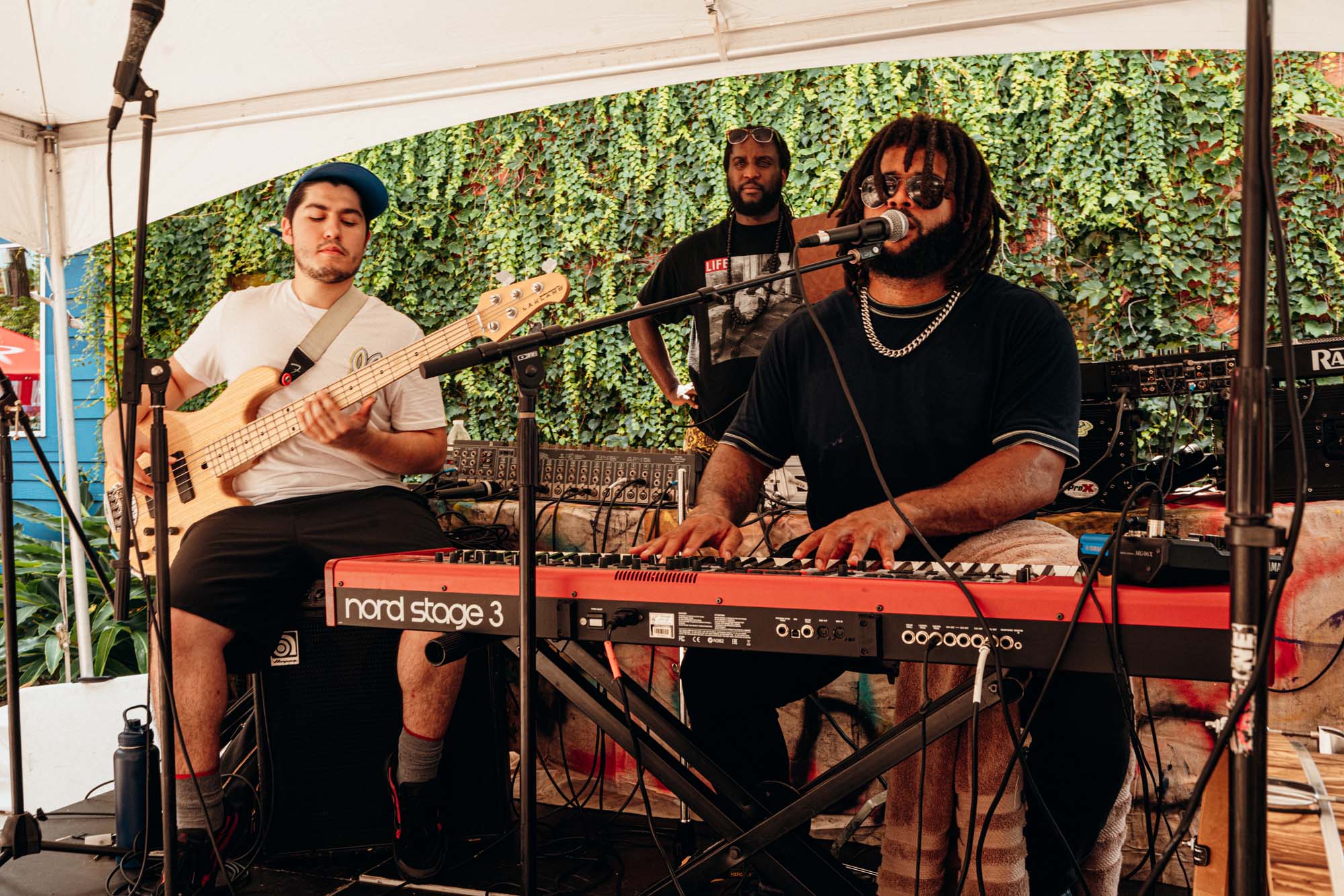 Brandon, wearing a black T-shirt and dark Aviator style sunglasses with medium-length dreaded hair, sits at a keyboard playing and singing. Another man, in a white T-shirt and sideways cap, stands behind him with a guitar.