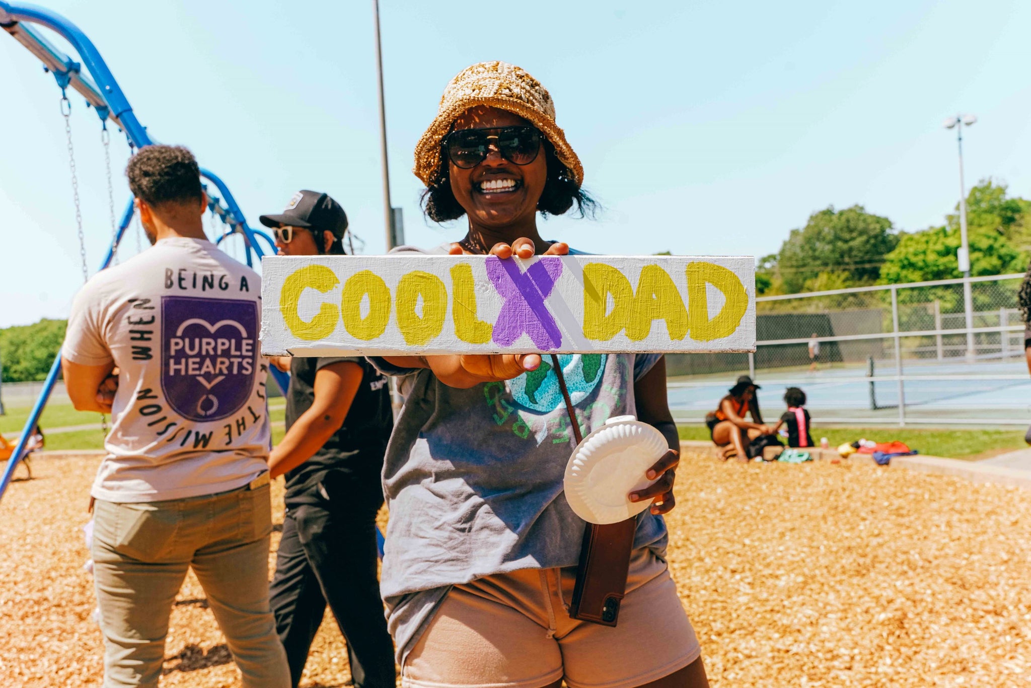 Danielle holds the wooden CoolxDad sign, hand-painted by kids. She has short black hair underneath straw sun hat, folded up in front, and a gray T-shirt and smiling happily. Behind her is a playground and some dads standing next to each other. It's a beautiful sunny day.