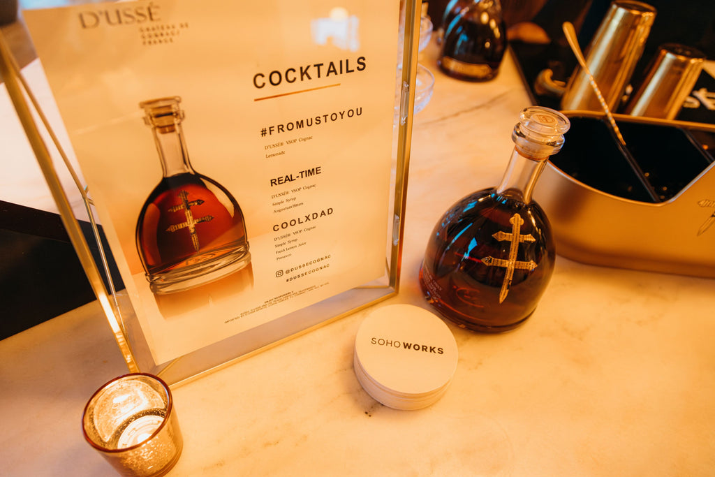 An overhead shot of a cocktail menu and a bottle of D'usse. The menu features cocktails called "Real-Time," Cool x Dad," and "#FromUsToYou." A pile of white coasters have black text in their center that reads: Soho Works.