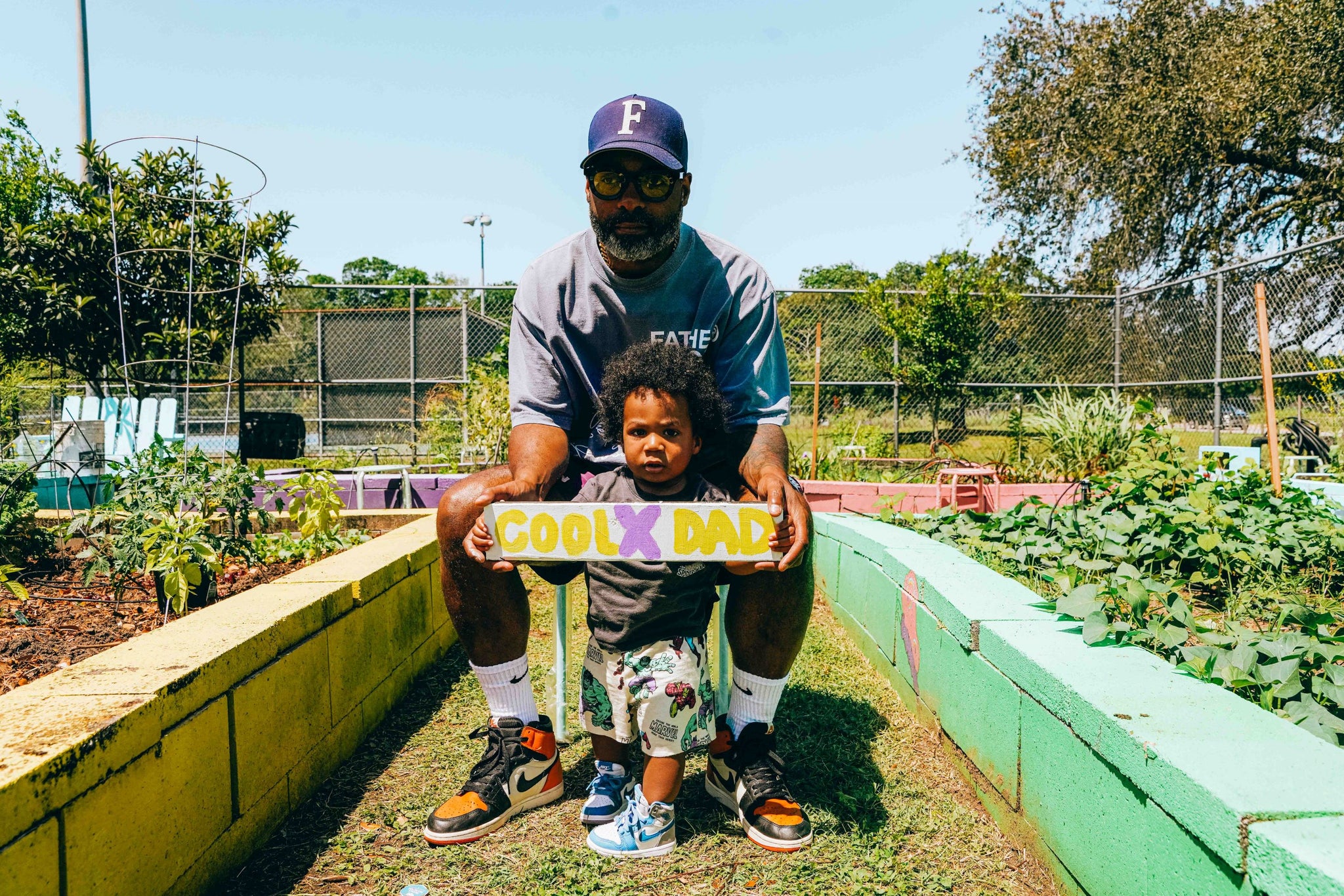Kevin and his son Lenox standing between rows of garden beds. Lenox, who is a toddler with curly black hair, is holding a wooden sign that says CoolxDad. Kevin is crouching behind him holding the sign's edges. He's wearing a T-shirt and a baseball cap and looks serious. It's a sunny day.