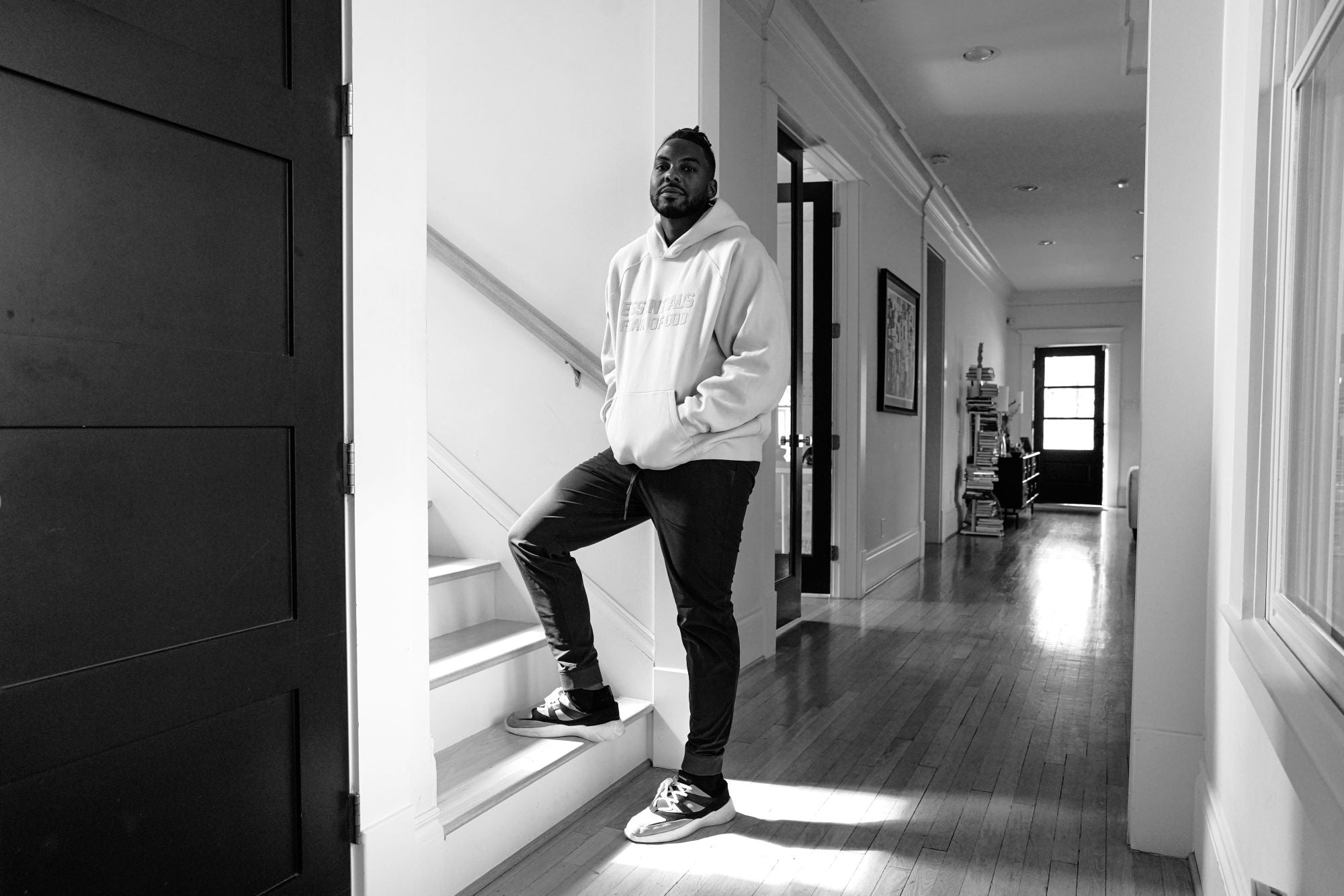 A black and white photo of Troy Hamm in his home. He's standing with one foot on the staircase, wearing black jeans and a white hoodie, and looking at the camera with a soft smile. Behind him is a hallway.