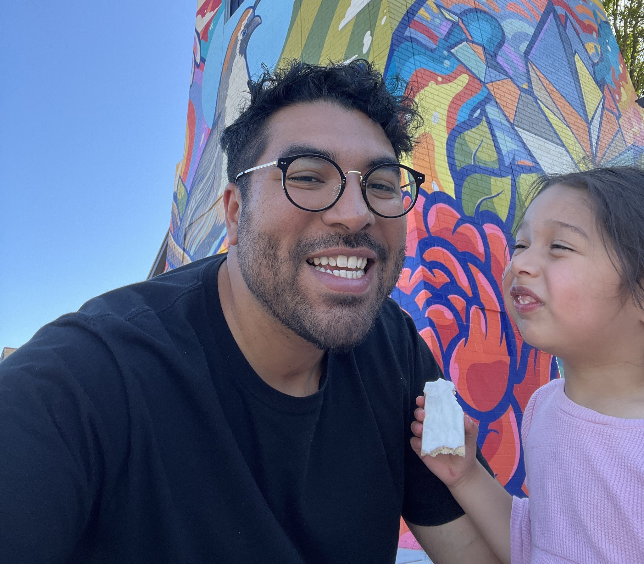 Anthony, smiling in a black T-shirt, with his daughter, who is eating something. Behind them is a colorful mural.