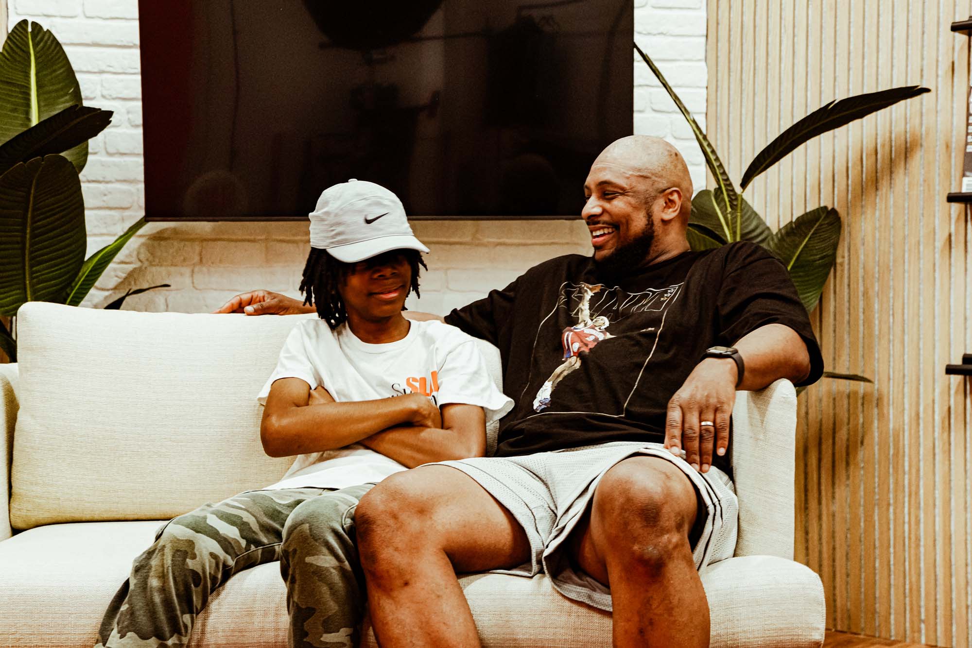 Les and his son on the couch in the slam office. Les is bopping his son's hat down and they're both laughing.