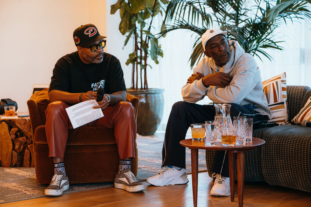 Two men sitting on chairs leaning over their knees. The man on the left, Kevin, is wearing a black CoolxDad T-shirt, burgundy pants, a black ball cap, and gray sneakers. To his right, the man is wearing a white ball cap and a gray sweatshirt. They're both looking attentively at some speaking off-camera.