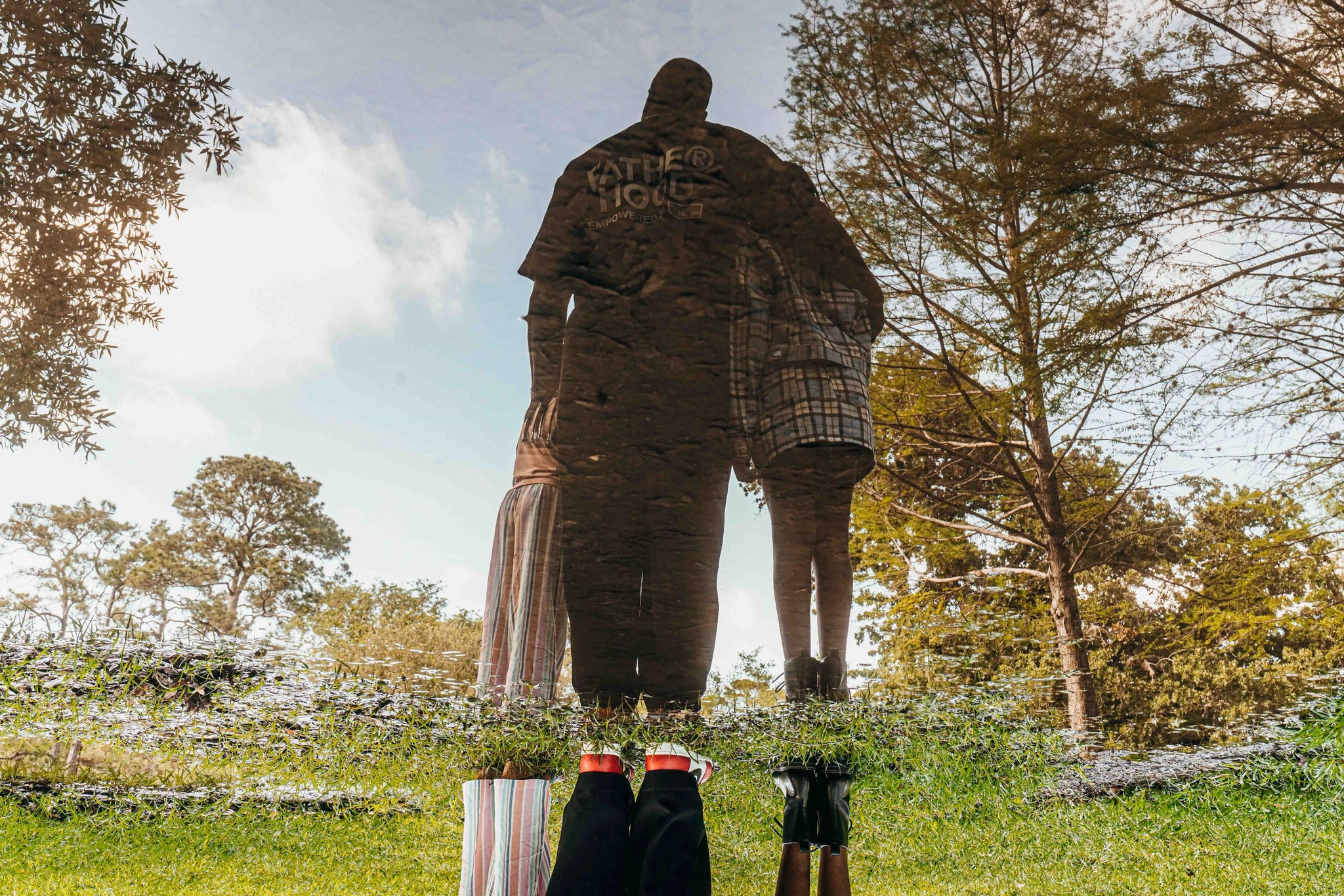 The reflection of Fred and his two daughters in a body of water. In the reflection, the family has their backs facing us. Fred has his arms around his daughters. at the bottom of the image, the family's real feet meet their reflections. Fred is wearing red and white Nikes and his daughters wear combat boots. 