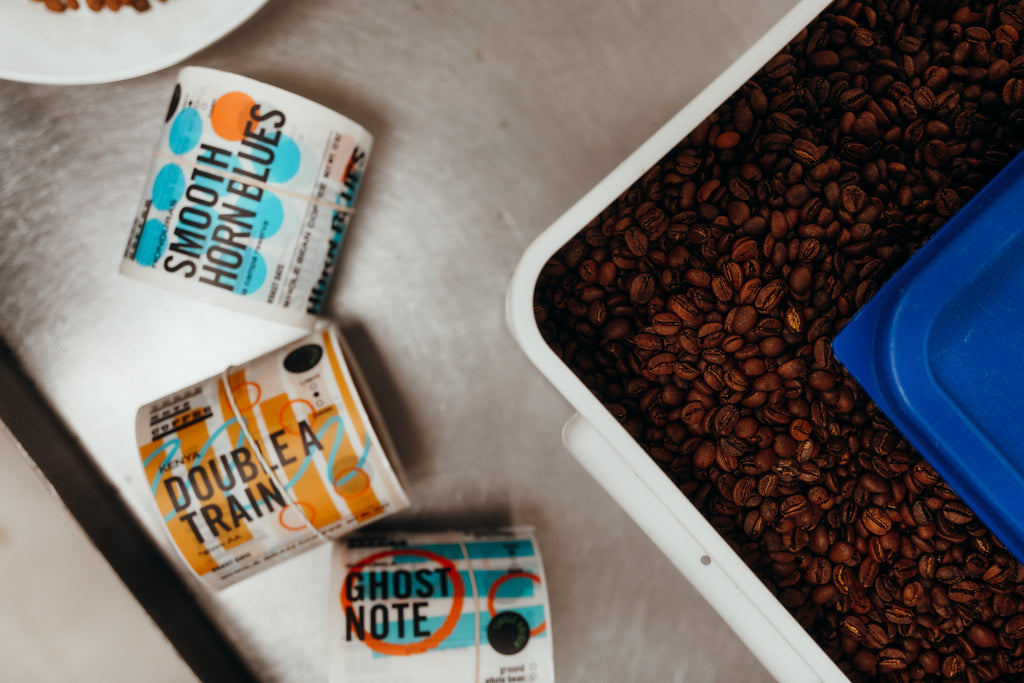 A corner of a tin of coffee beans next to a roll of stickers featuring the coffee flavor names.