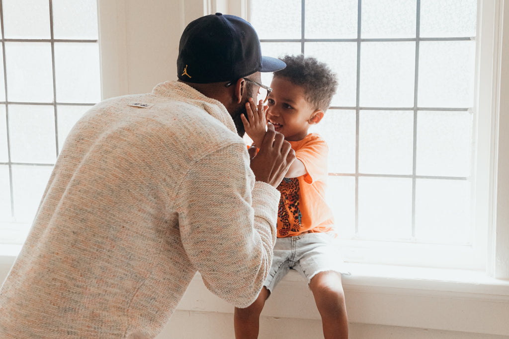 Tio's toddler son sits on the windowsill, smiling and holding his hands out to his father's face.