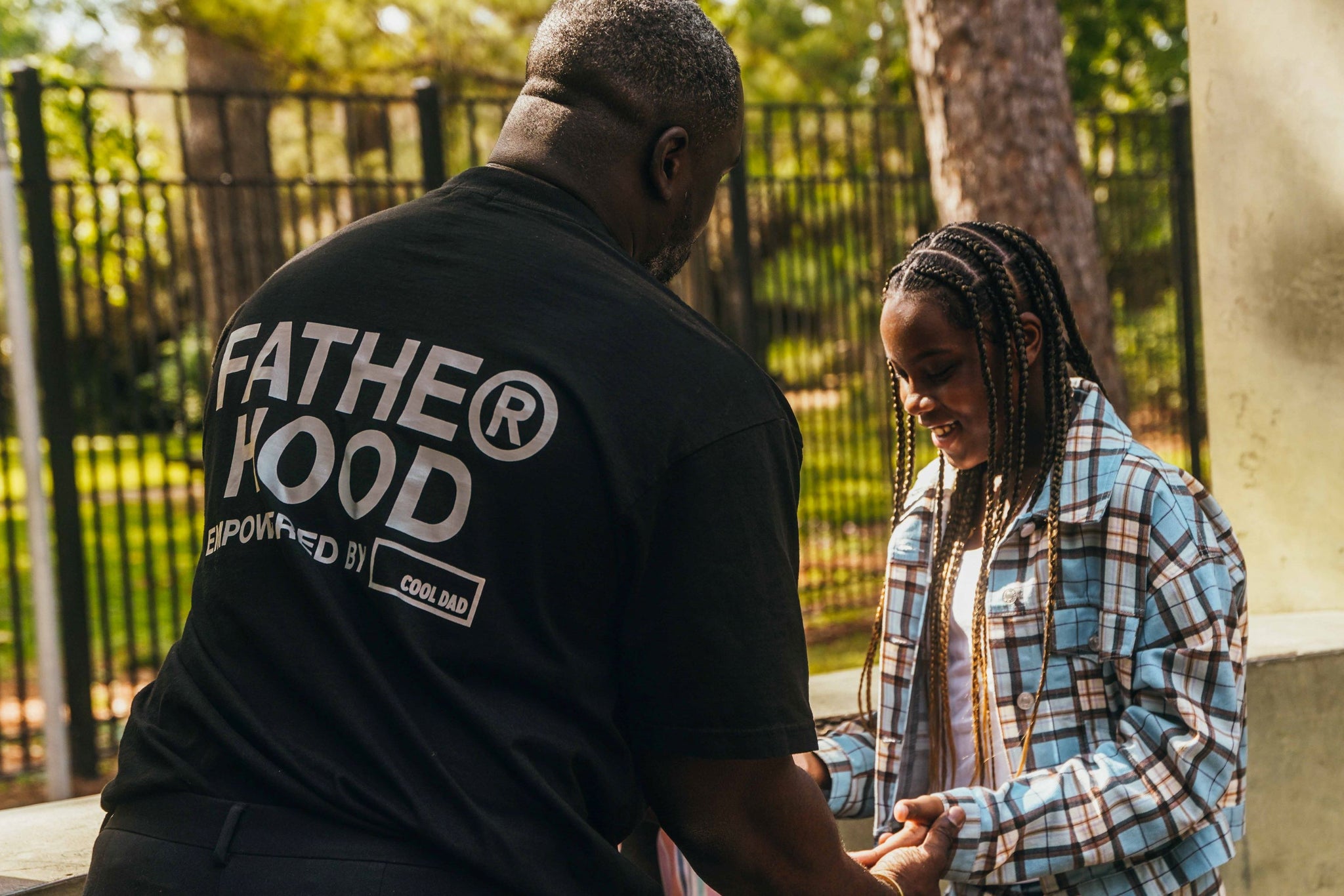 The back of Fred's torso. He's wearing a shirt that says "FATHER HOOD" with the r in the symbol of the registered trademark sign. He has his hands out toward his daughter, who has on a blue checkered shirt and is wearing braids. They appear to be playing a hand game, like paddycake. She's smiling.
