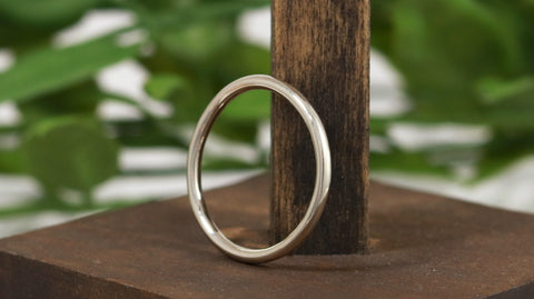 A 2mm thick white gold ring is leaning against a piece of dark wood with a background of greenery. The ring is made from solid rounded 14k white gold.