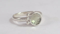 A round rose cut green amethyst stone is set in a sterling silver bezel on a sterling silver ring band. There are three silver bubbles along one side of the stone. 