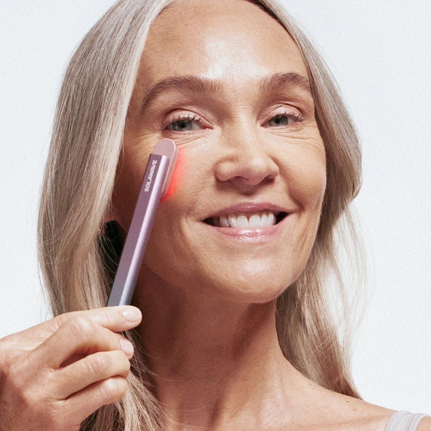 Woman using a red light therapy device near her eye, smiling.