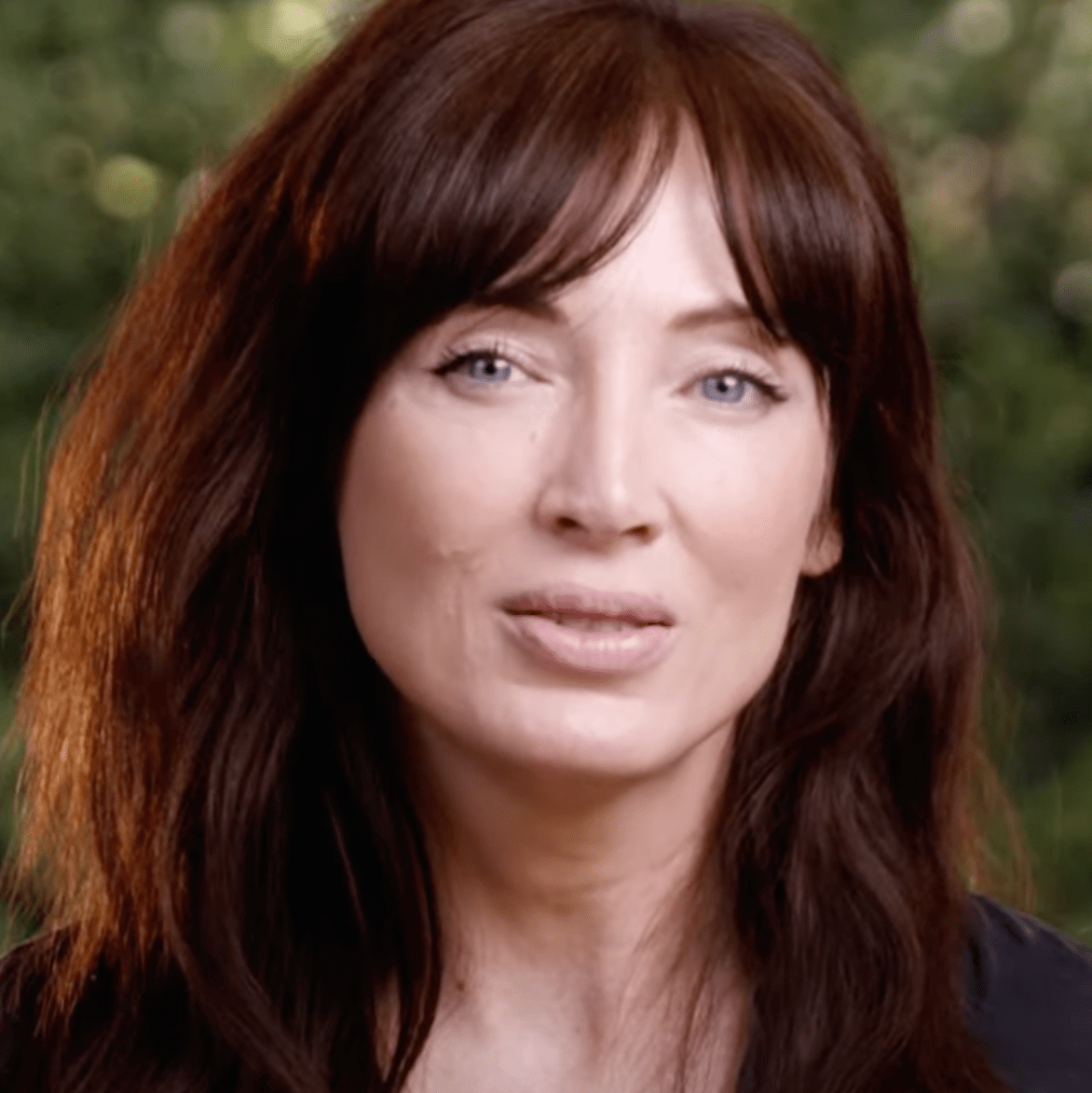 Woman with long brown hair and bangs in front of a blurred green background.