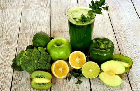 A fresh green juice surrounded by its whole ingredients: green apples, lime, lemon, kiwi, broccoli, and bell pepper