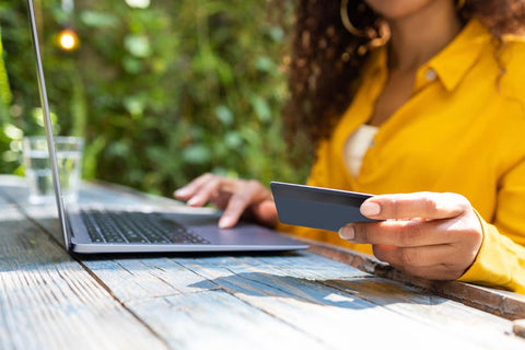 A person in a yellow blouse holds a credit card while typing on a laptop
