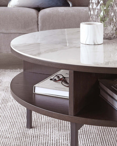 Round coffee table with a white marble top and lower dark wooden shelf, featuring a textured surface, partially resting on a light-coloured carpet, with a grey sofa in the background. The table is adorned with a decorative glass vase and a smaller, cylindrical white marble container, and holds a book and a pair of eyeglasses underneath.