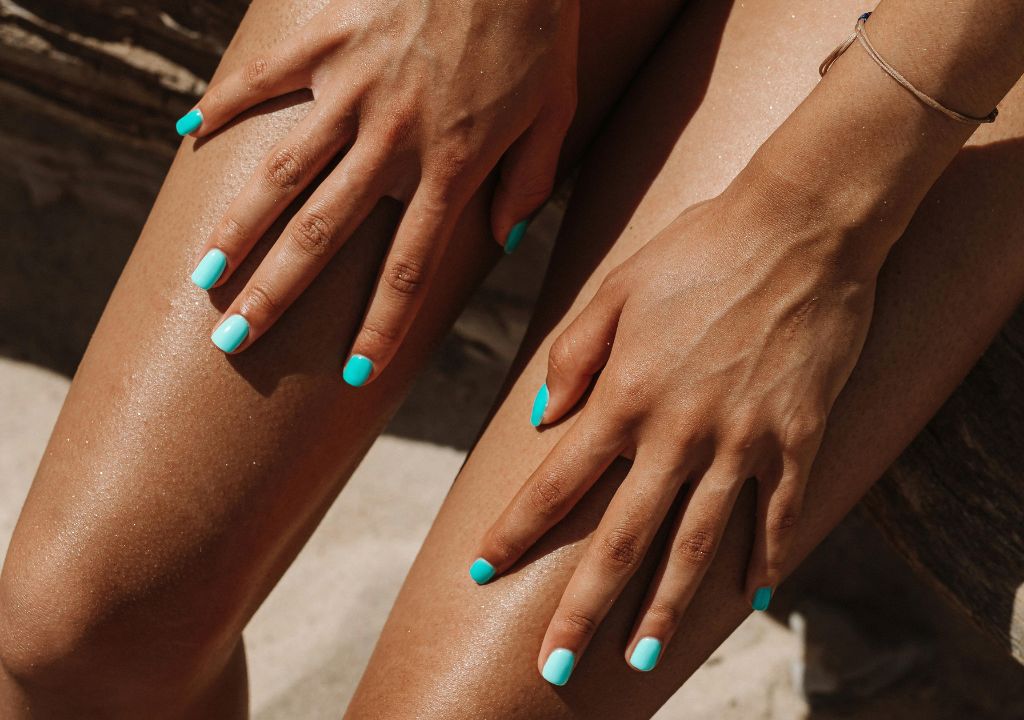 tanned hans with blue nail polish applying lotion to legs