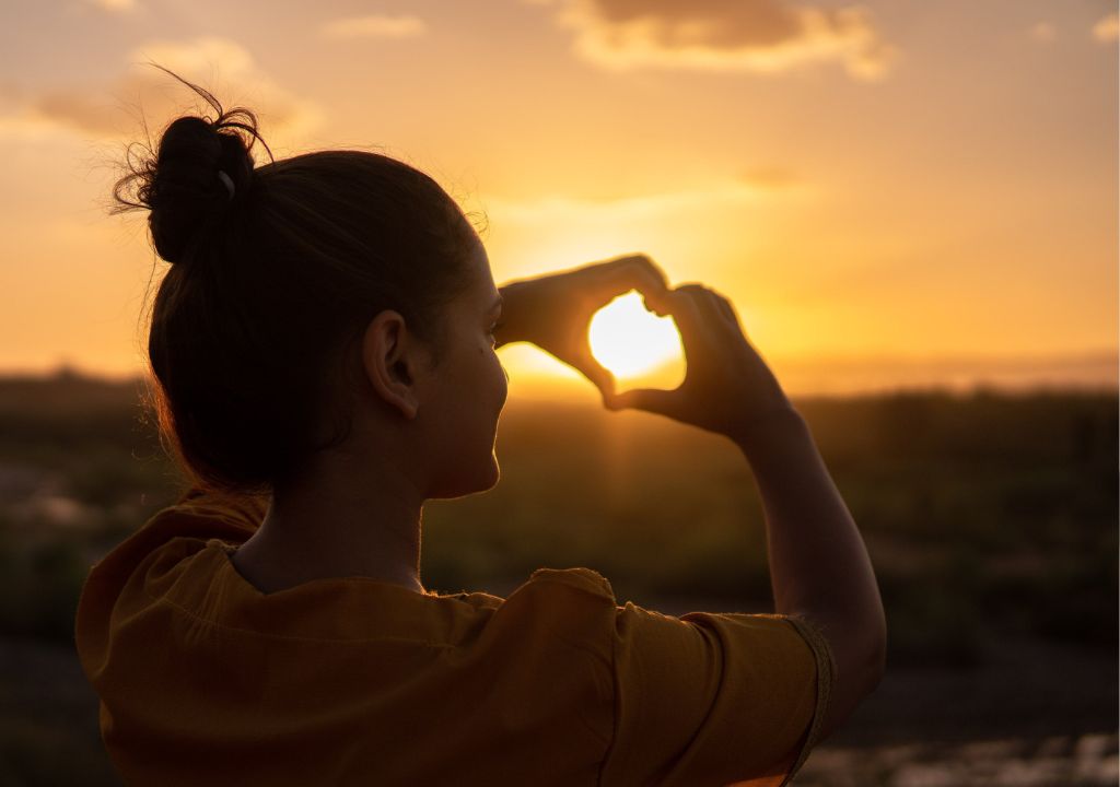 lady holding her hands to create a heart shape against the sunset