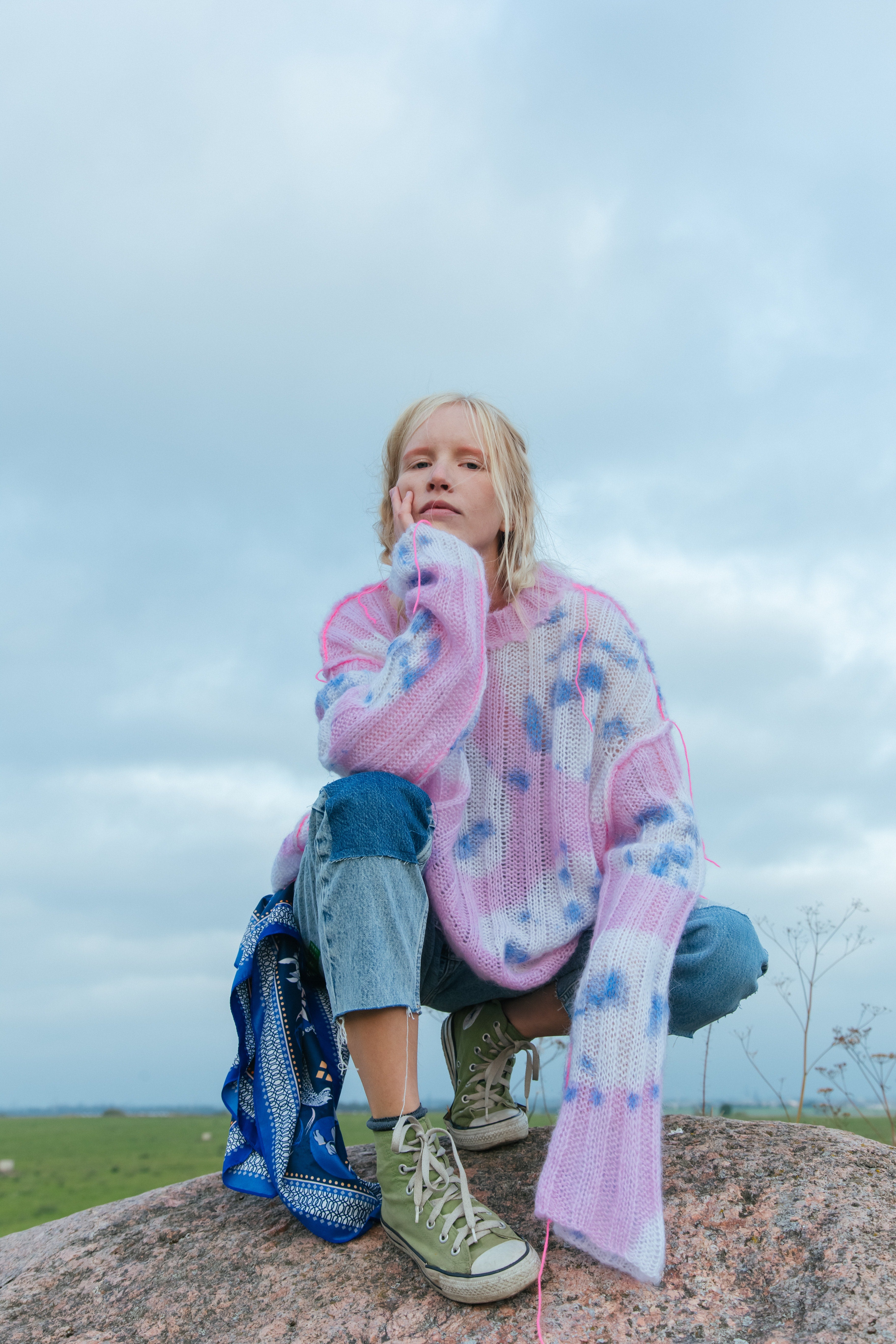 A girl in jeans, green sneakers and a pink spotted sweater sits on a stone