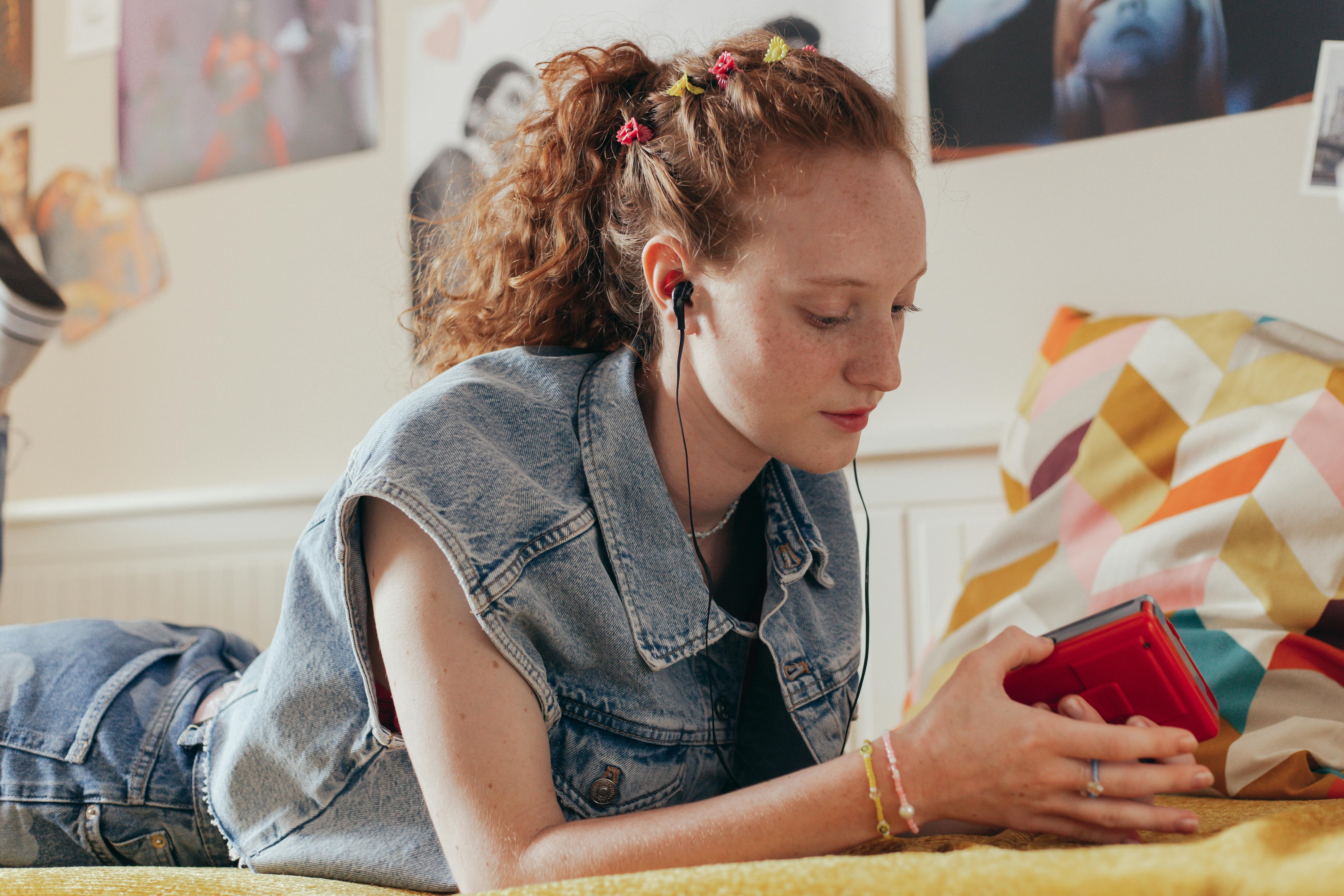 A girl in a denim vest and with headphones in her ears lies on the bed