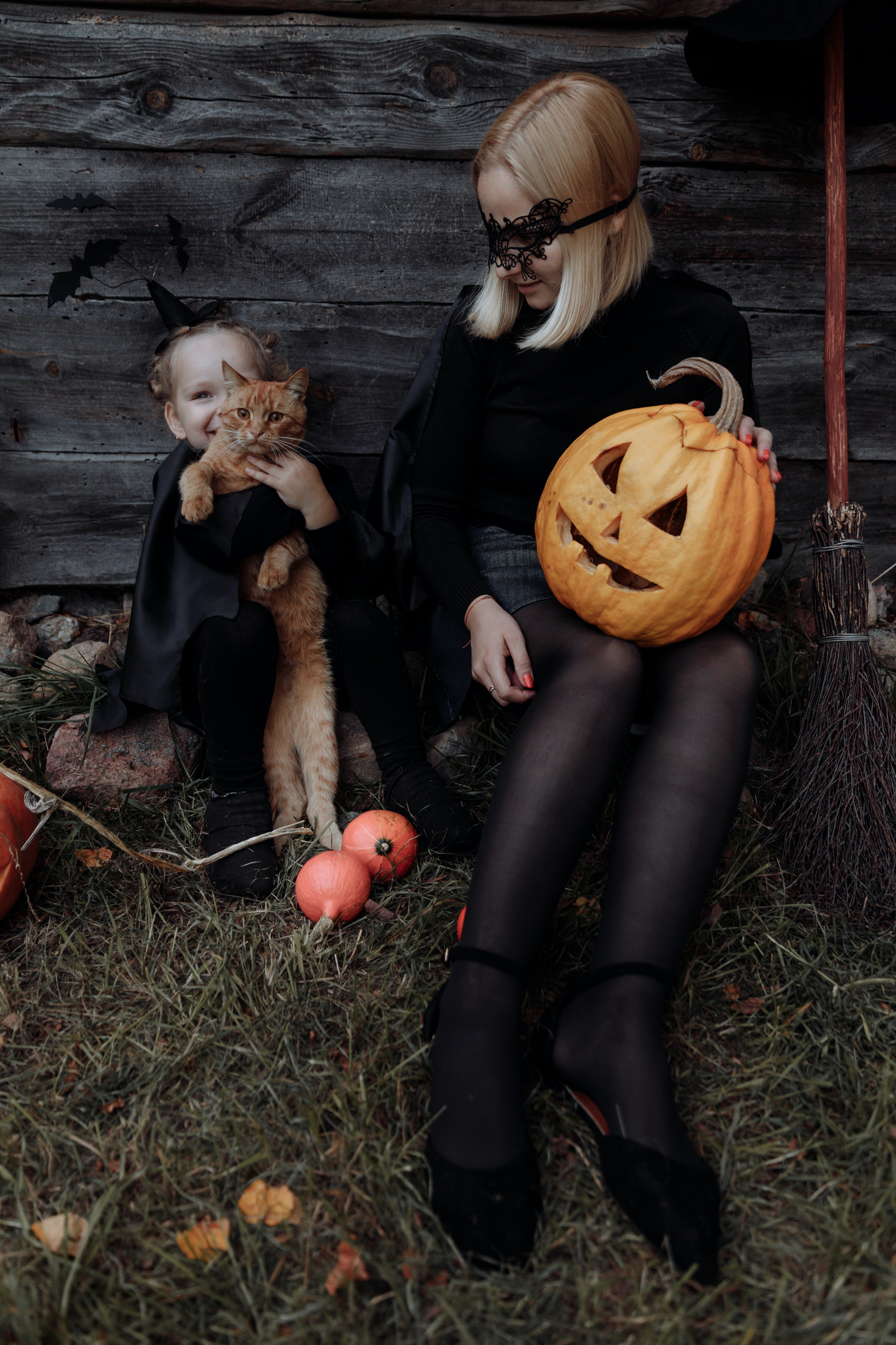  A woman is holding a jack-o'-lantern, a girl is holding a cat, both are dressed in witch costumes