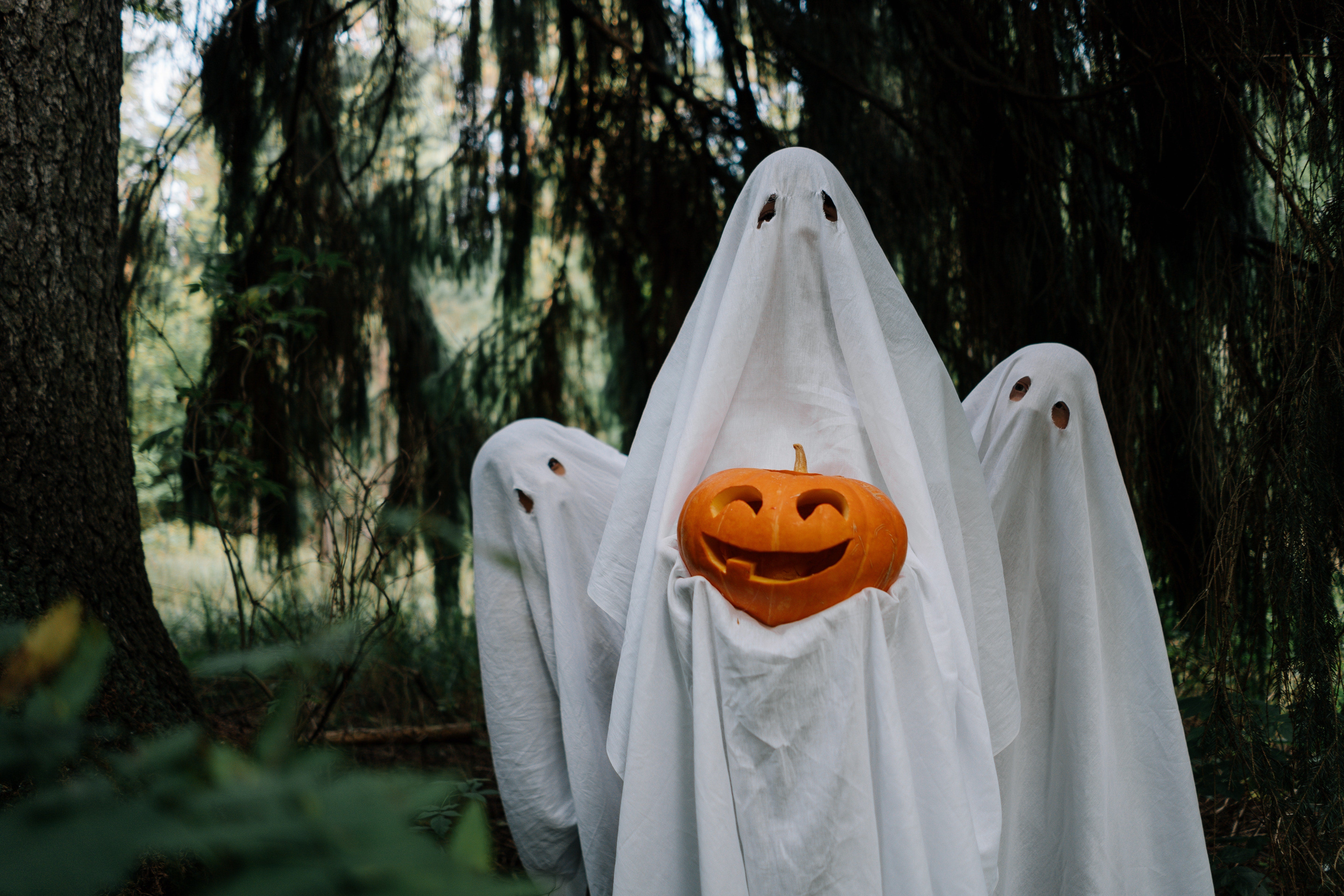 A man in a ghost costume holds a jack-o-lantern, behind him are two more people in ghost costumes