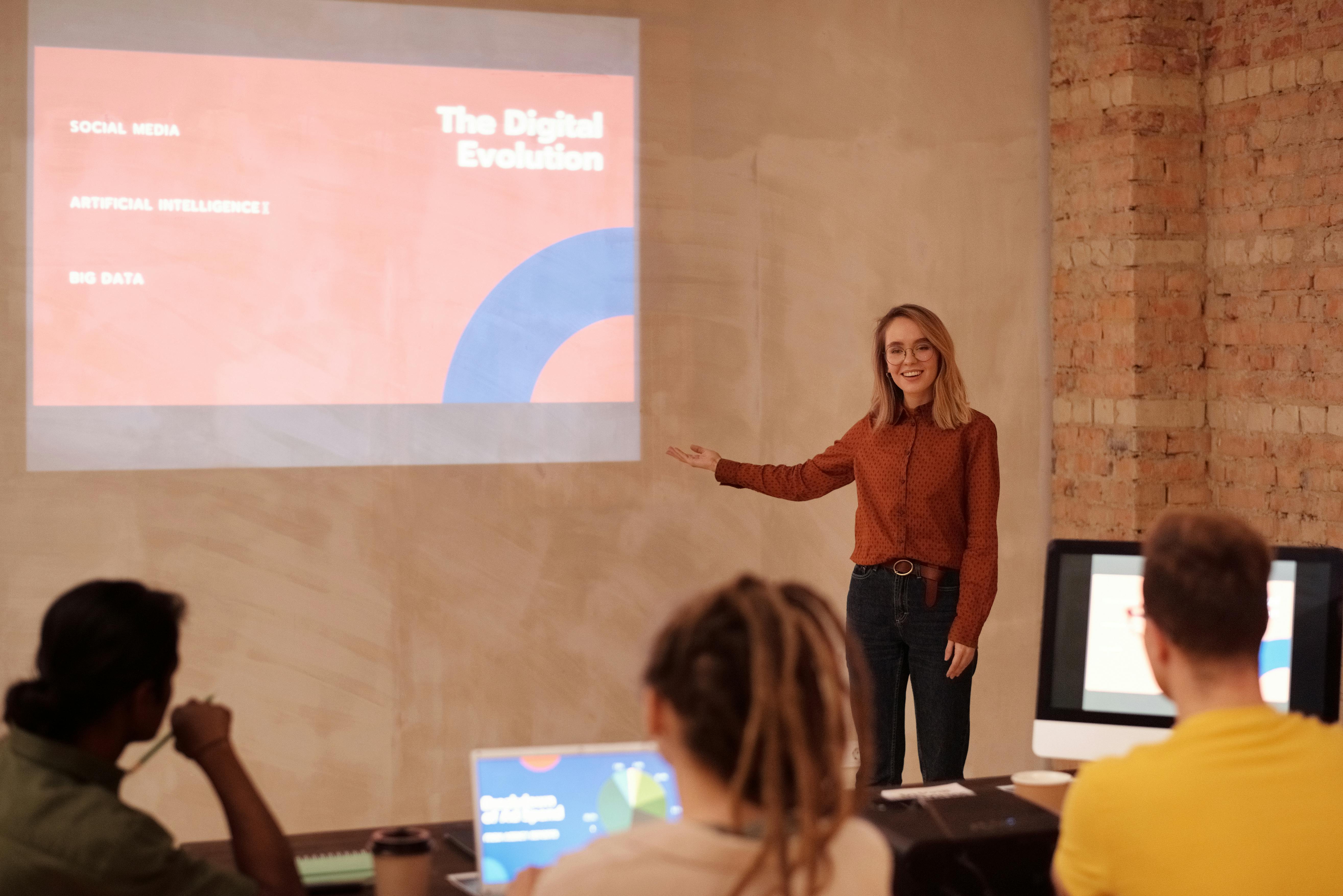 A woman speaks in front of an audience and points to an image from a projector