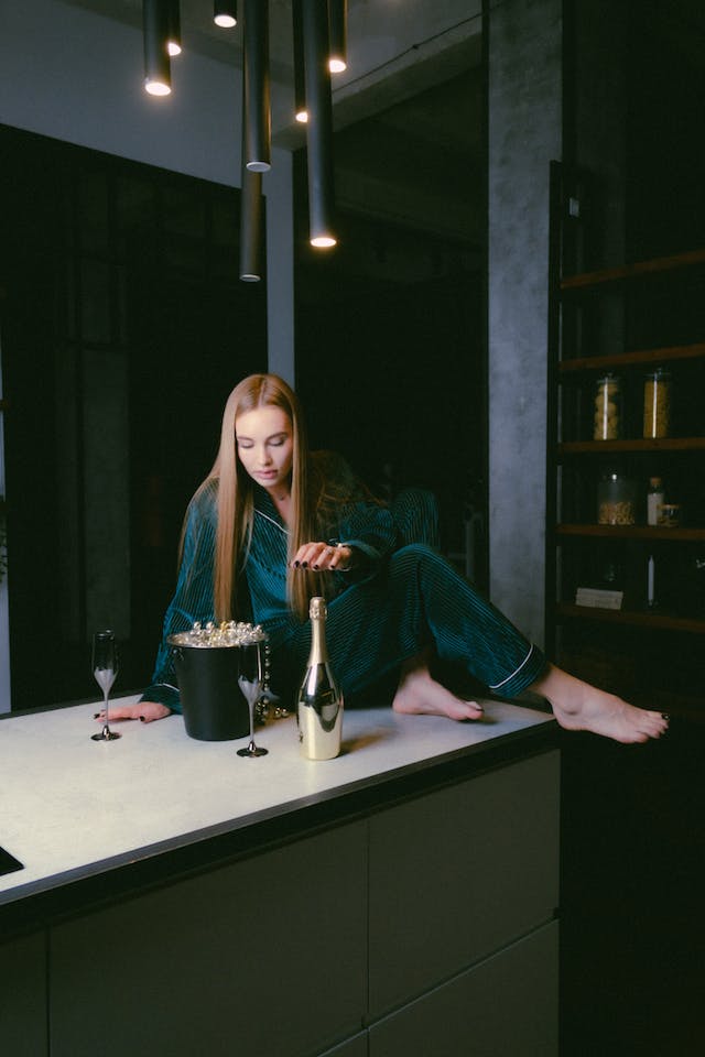 A girl in pajamas sits on the counter in the kitchen with a bottle of champagne and two glasses