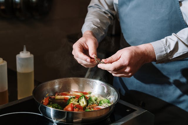 A man in an apron puts seasoning in a frying pan on the tile