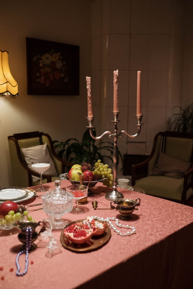 There are a bowl of fruit, plates, glasses and a candlestick with three candles on the table