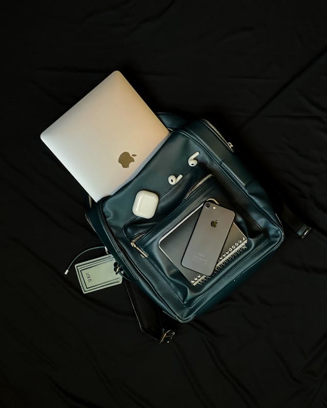 A laptop sticks out of the bag, a notepad, a smartphone and a headphone case are on top