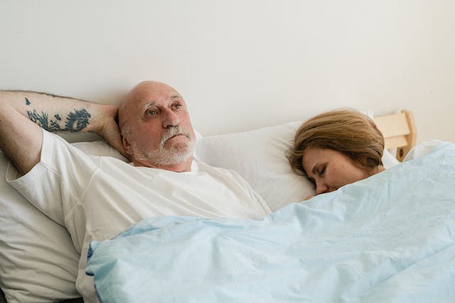 An elderly man lies on the bed with his hand behind his head, next to him is a woman