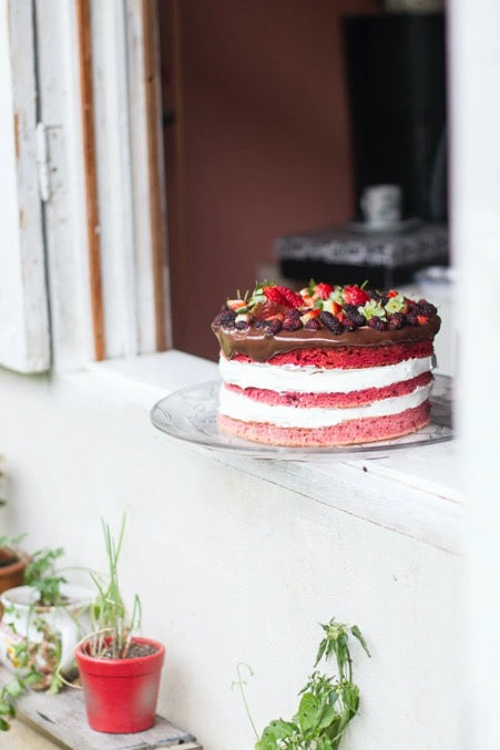 The cake stands on a white window sill, and there are flowers in pots below