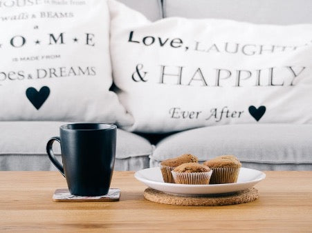 There is a black mug on the table, a white plate with muffins, and in the background a gray sofa with white pillows