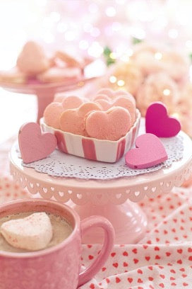 There is a pink mug on the table, a pink stand with a box containing heart-shaped cookies