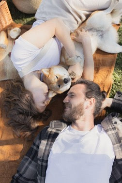  A woman and a man lie on a blanket on the grass, a woman hugs a dog