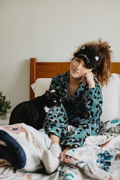 A black cat sits next to a girl in pajamas wearing a cat head mask