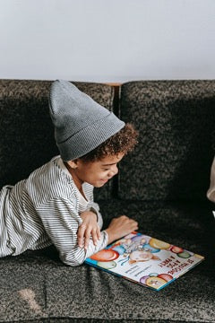 A child in a striped shirt and a gray hat lies on the sofa and looks at a book