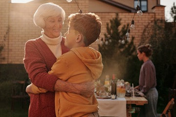 An elderly woman in a red sweater hugs a boy in an orange sweatshirt on the background of a brick house