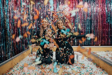 Two girls in black clothes throw colored pieces of paper into the air