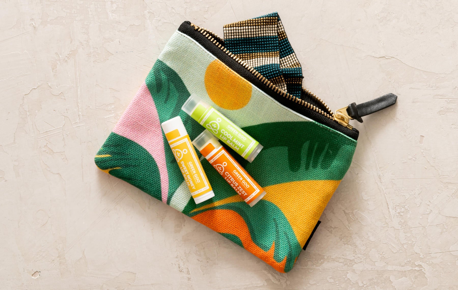 Green Goo's Citrus Zest, Cool Mint, and Sweet Honey lip balms laying on a tropically floral makeup pouch