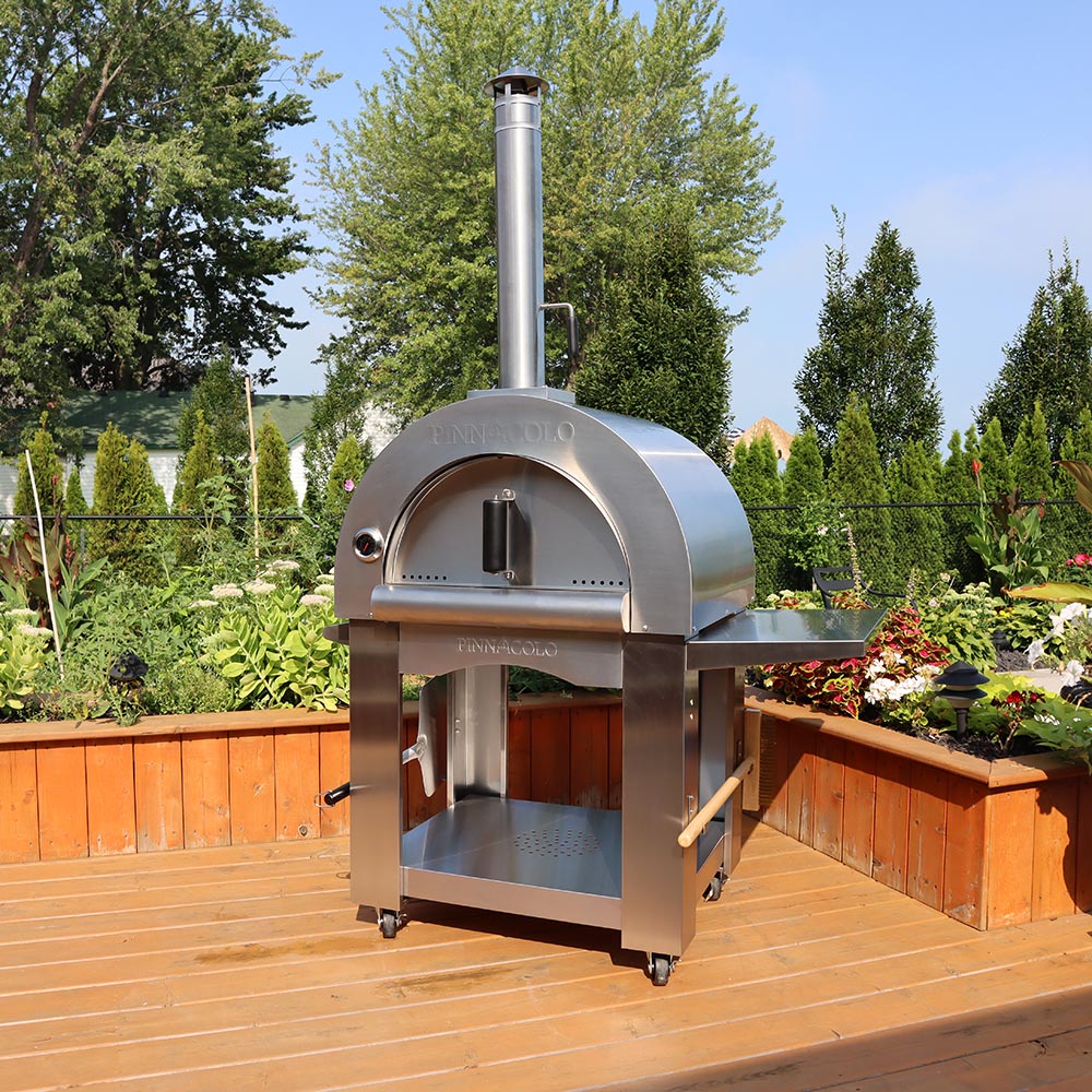 Metal pizza oven sitting on a wood patio with a garden in the background.