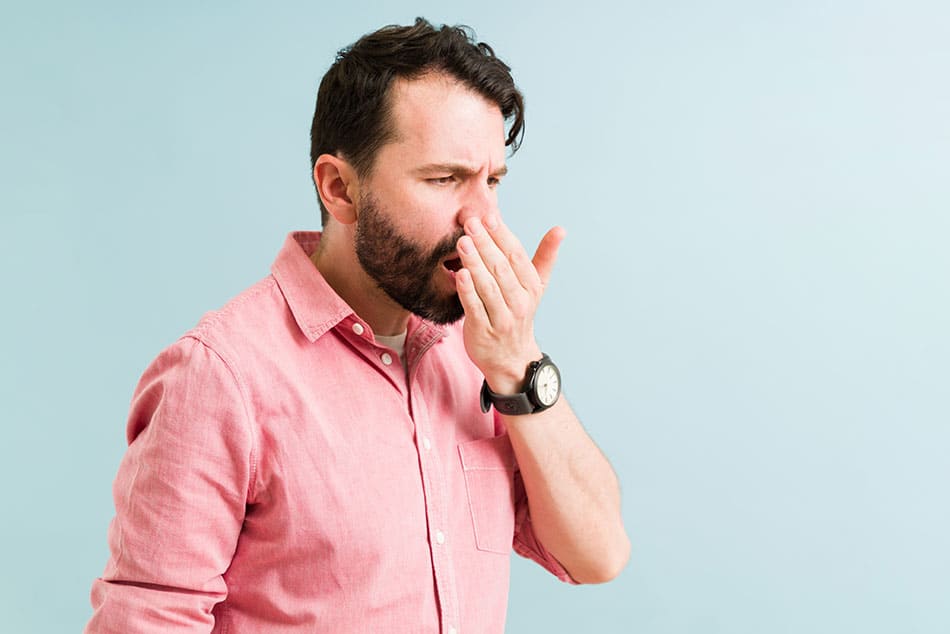 Man stood up breathing on his hand with a thoughtful look on his face, in front of a white and blue gradient background.