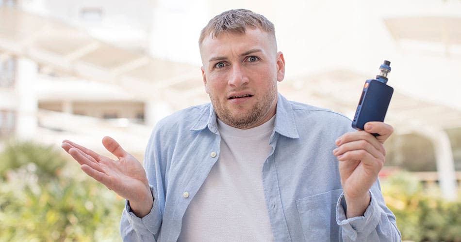 Man holding a vape kit in one hand with shrugged shoulders with a confused face.