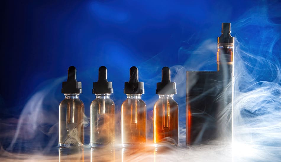 Four bottles of dark brown e-liquid next to a vape kit with vapour surrounding them.