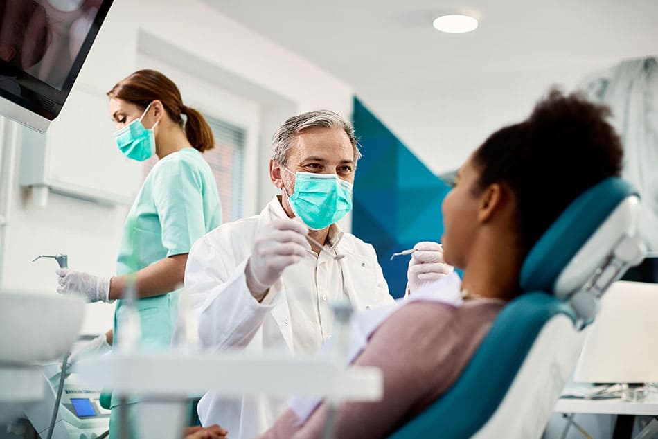 Dentist looking towards a woman sat in chair with a hygienist in background preparing utensils.