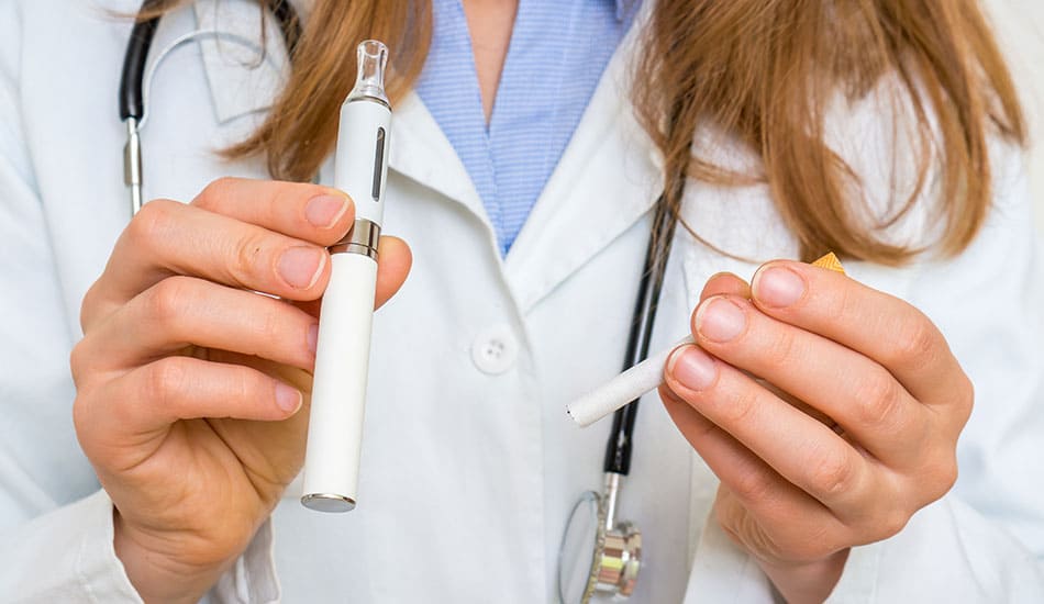 Doctor in white gown holding up a vape kit in one hand and a cigarette in the other hand.