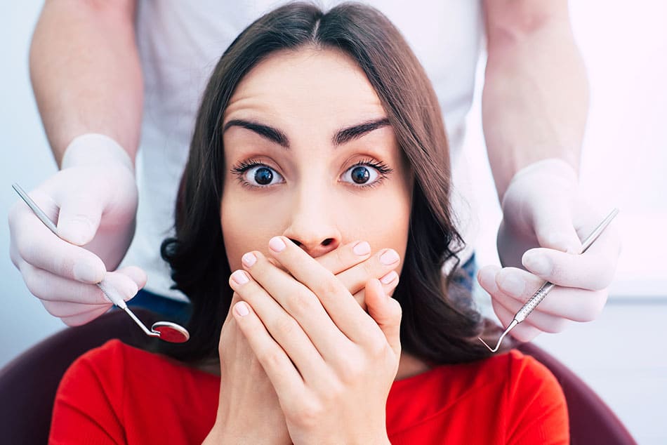 Woman looking towards camera with hands over her mouth with a dentist holding utensils either side of her head.