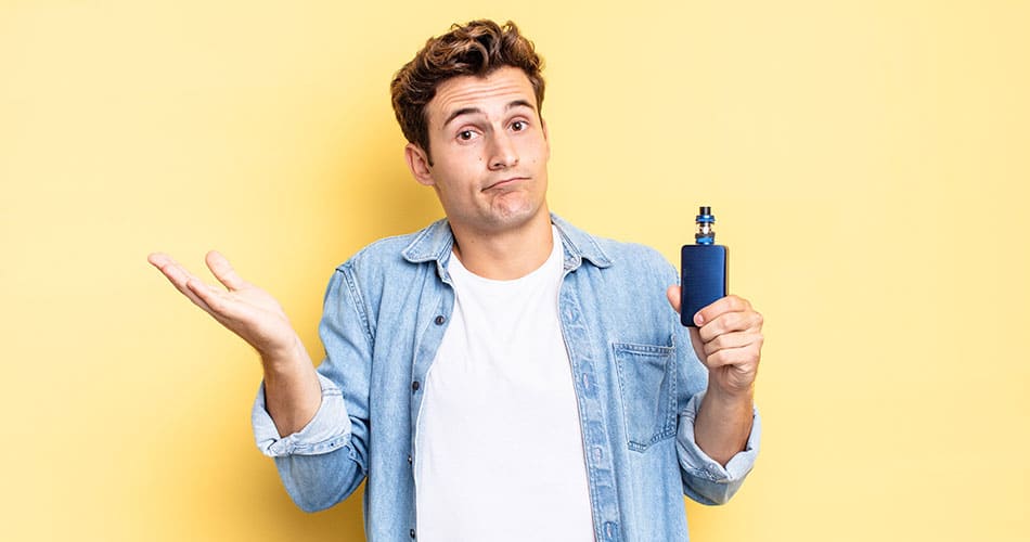 Man holding a vape in one hand and raised empty hand with shrugged shoulders and a thinking expression on his face, stood against a bright yellow background.