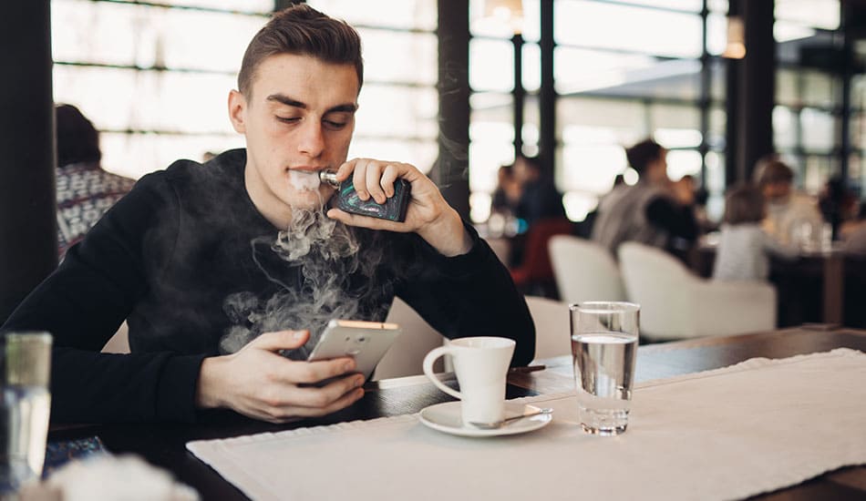Man sat at restaurant table reading his phone while vaping an e-cigarette.