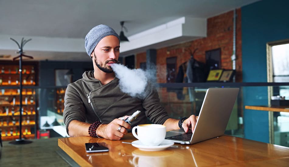 Man sat at a table working on his laptop whilst vaping an electronic cigarette.